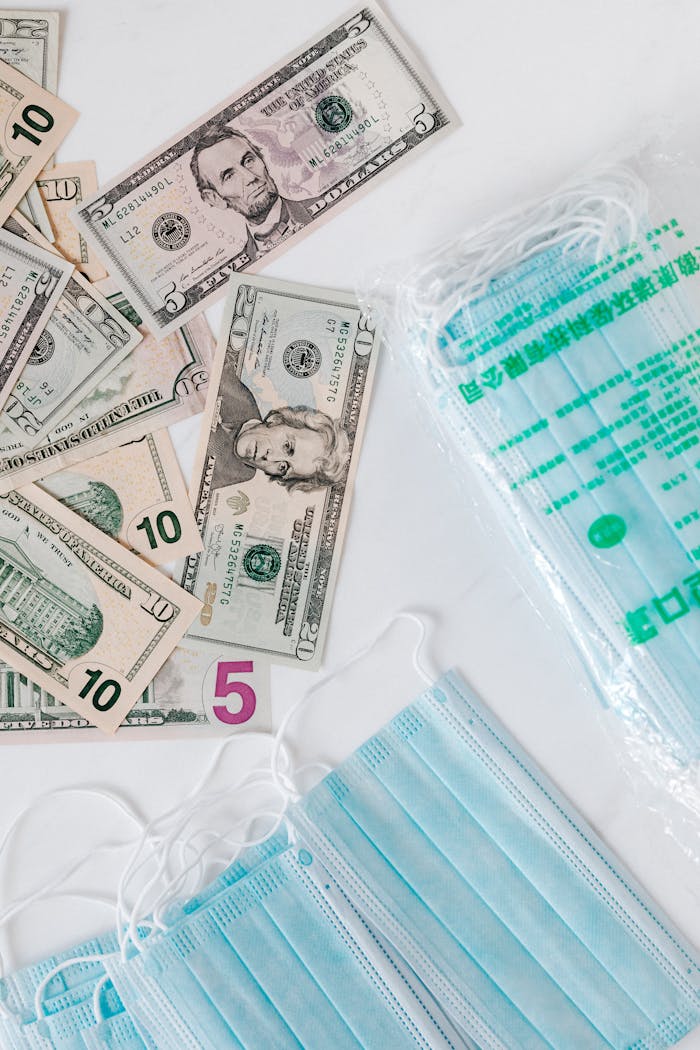 services-01 Top view composition of disposable medical masks placed on white table near scattered randomly various paper banknotes of American dollars